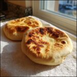 Two freshly baked, puffy pita bread rounds cooling on a white towel near a sunlit window.
