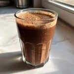 A tall, faceted glass filled with rich, dark chocolate milk topped with a layer of foam.