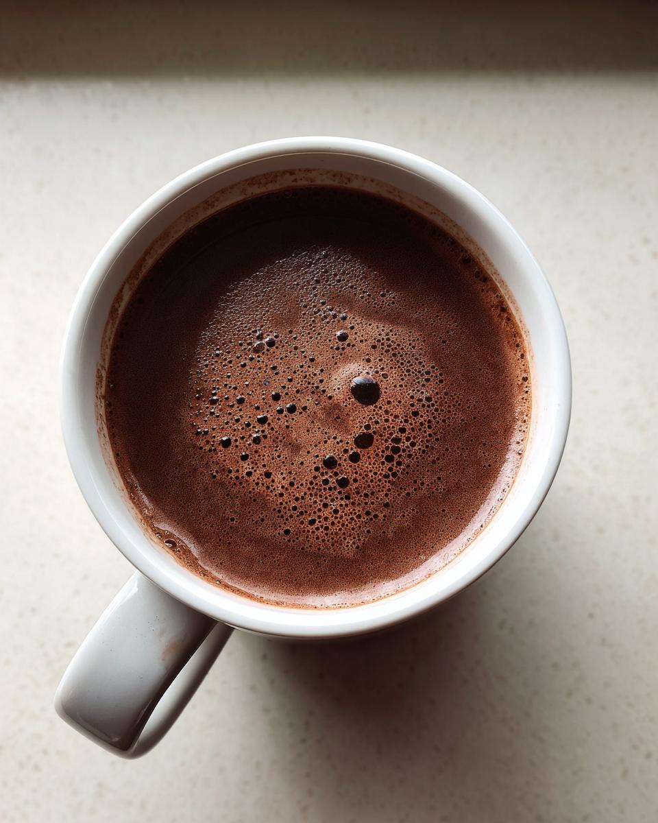 Overhead view of rich, dark brown hot chocolate with small bubbles on top, served in a white mug.