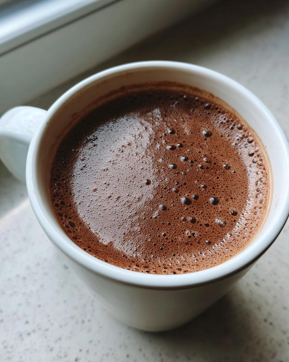 Close-up of rich, dark brown hot chocolate with a foamy top in a white ceramic mug.