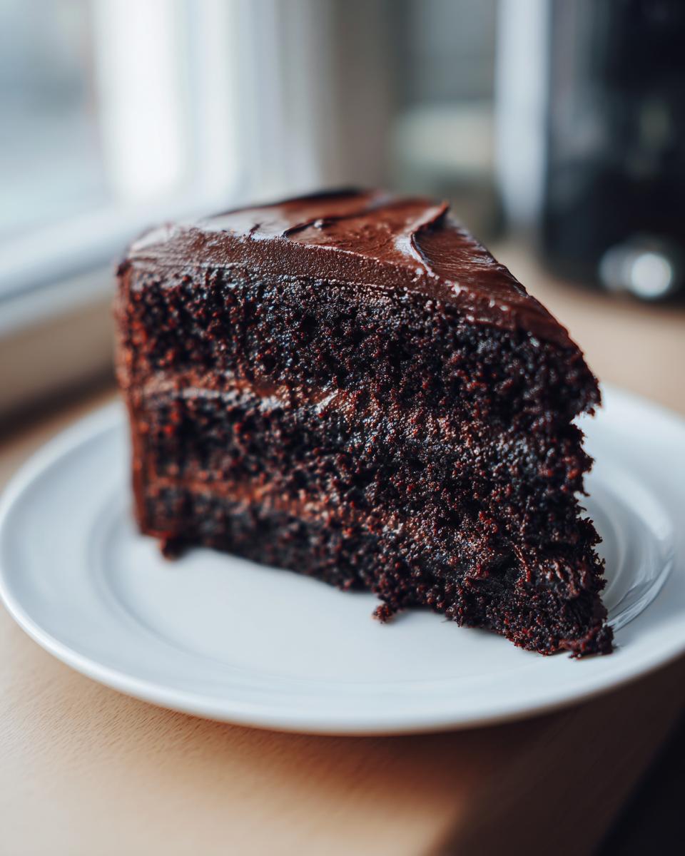 A close-up of a rich, dark slice of moist chocolate cake layered with thick chocolate frosting on a white plate.