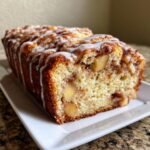 A freshly baked apple fritter bread loaf drizzled with white icing, showing chunks of apple inside.