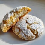 Two gooey butter cookies, one broken in half to show the soft, creamy center, dusted heavily with powdered sugar.