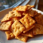 A close-up, appetizing photo of a pile of golden, square homemade cheese crackers seasoned with visible specks of pepper.
