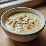 A close-up of a white bowl filled with rich, creamy cream of chicken soup, featuring shredded chicken, carrots, and celery.