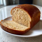 A loaf of homemade whole wheat bread with one thick slice cut and resting beside it on a white plate.
