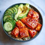 Close-up of Honey Sriracha salmon bowls featuring glazed salmon chunks over rice with sliced avocado and cucumber.
