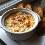 A bubbling, hot baked reuben dip topped with melted cheese and ham pieces, served with toasted bread for dipping.