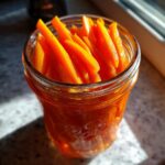 Close-up of bright orange, julienned pickled carrots filling a glass mason jar, catching sunlight.