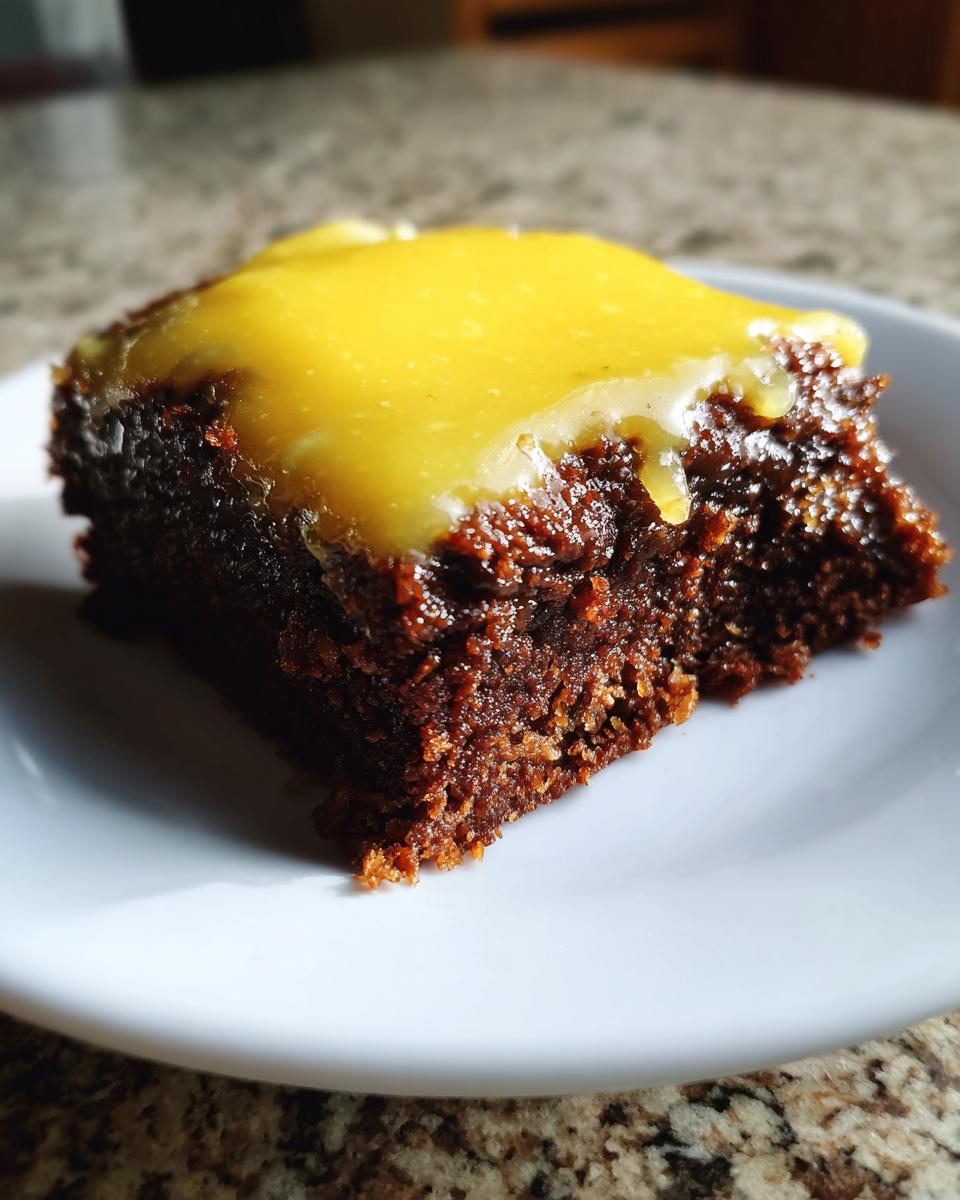 A close-up of a rich, dark slice of lemon brownies topped with a thick, bright yellow lemon glaze.
