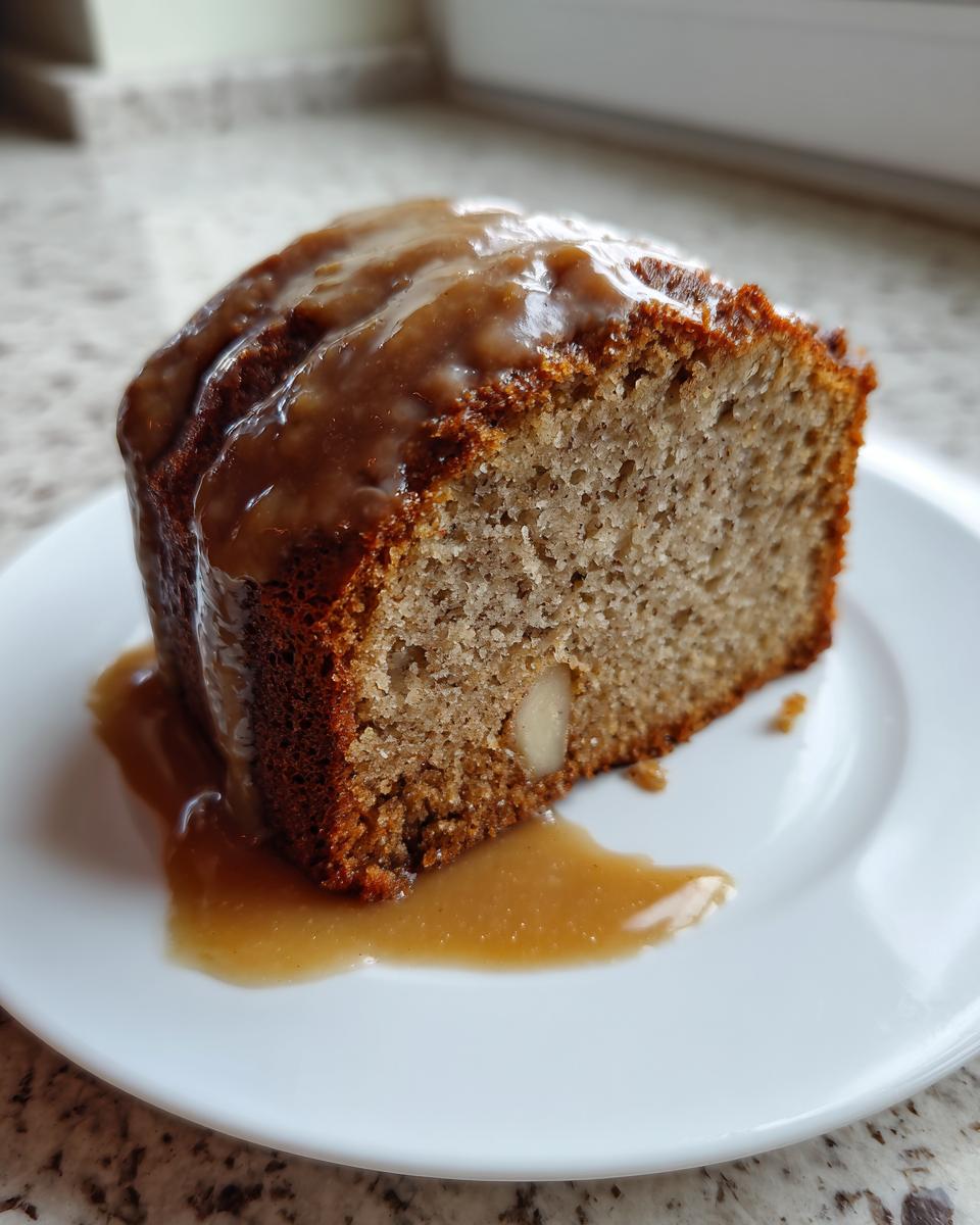 A close-up of a moist slice of apple dapple cake topped with a rich caramel glaze, served on a white plate.