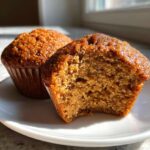 Close-up of two moist bran muffins on a white plate, one having a bite taken out to show the texture.
