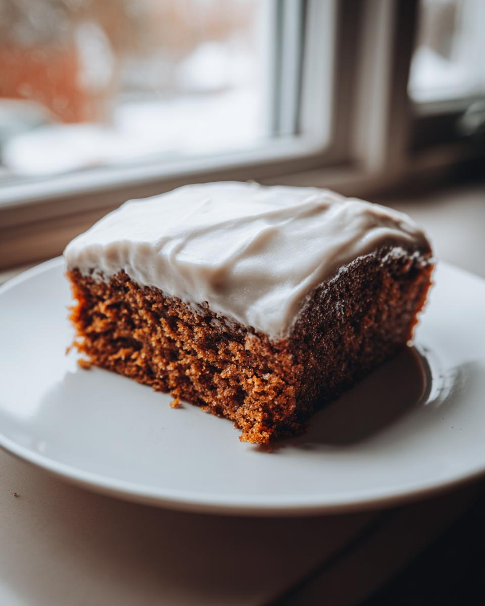 A close-up of a moist slice of gingerbread cake topped with thick white frosting, sitting on a white plate.
