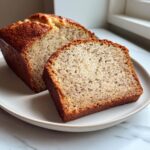 A slice of moist greek yogurt banana bread next to the rest of the loaf on a light plate.