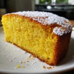 Close-up of a moist, bright yellow slice of olive oil cake dusted with powdered sugar on a white plate.
