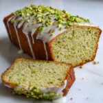 A loaf of moist pistachio bread, partially sliced, topped with white glaze and chopped pistachios.