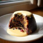 A rich, dark plum pudding with a slice removed, generously topped with creamy custard sauce.