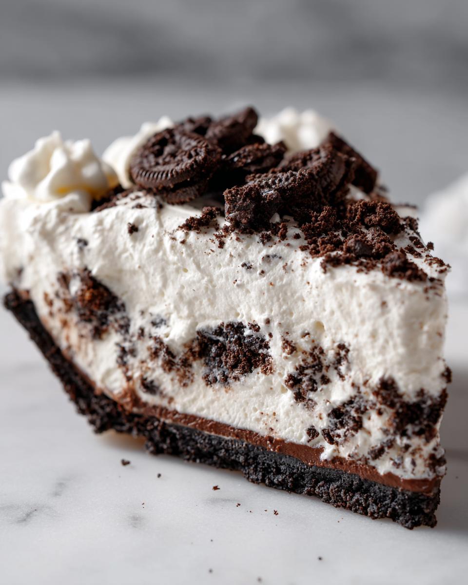 A close-up slice of rich no-bake Oreo pie featuring a dark crust, chocolate layer, and fluffy white filling topped with crushed Oreos.