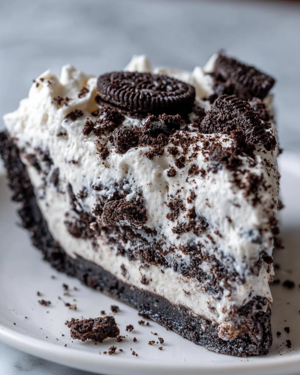 A close-up slice of rich, creamy no-bake Oreo pie with a dark crust, topped with whipped cream and cookie crumbles.