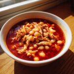 A close-up of a white bowl filled with vibrant red chicken pozole rojo, featuring shredded chicken and white hominy kernels.