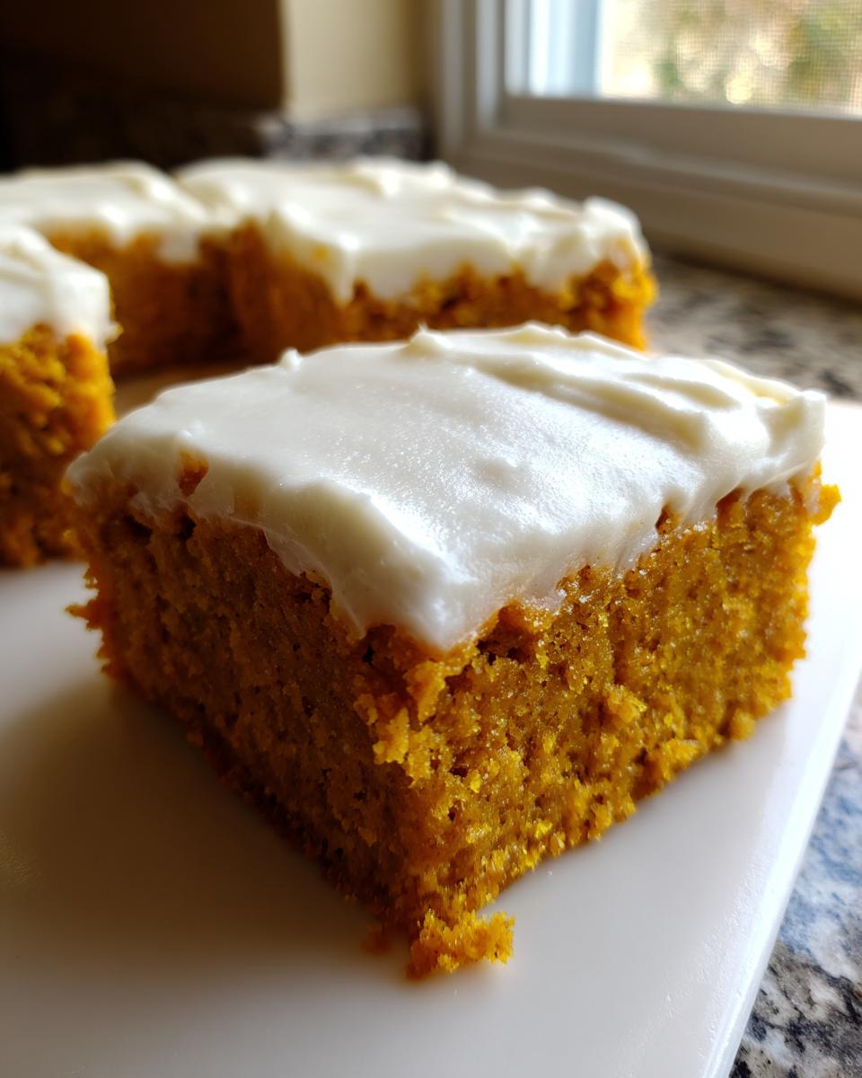 A close-up of a moist, orange pumpkin bar topped with thick, white cream cheese frosting.