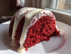 A close-up slice of vibrant red velvet bundt cake drizzled generously with white cream cheese glaze.