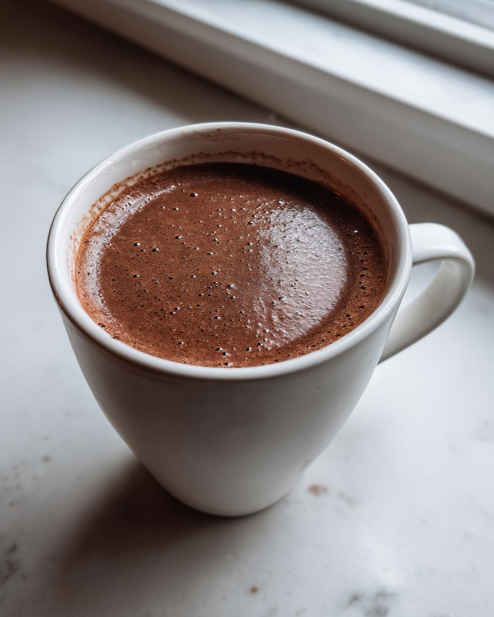 Close-up of rich, dark brown hot chocolate with small bubbles on top, served in a white mug.
