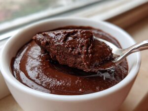 Close-up of a spoonful of rich, dark homemade chocolate pudding being lifted from a white bowl.