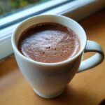 Close-up of a white mug filled with rich, frothy hot chocolate resting on a wooden surface near a window.