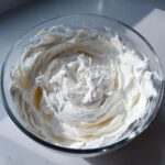 Close-up of thick, bright white, stiff-peak royal icing recipe mixture in a clear glass bowl.