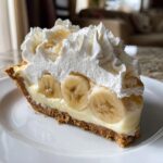 A close-up slice of rich banana cream pie featuring a graham cracker crust, vanilla custard, fresh banana slices, and a mountain of whipped cream.