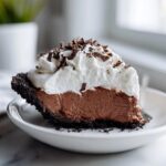 A decadent slice of chocolate mousse pie with a dark crust, topped with whipped cream and chocolate shavings.