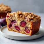 A thick slice of moist plum cake featuring baked-in red fruit halves and a generous brown sugar crumble topping.