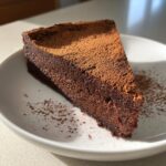 A dense, dark slice of flourless chocolate torte dusted generously with cocoa powder, served on a white plate.
