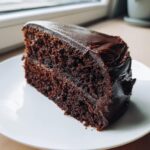 Close-up of a rich, dark slice of moist chocolate cake covered in thick, fudgy chocolate frosting.