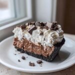 A decadent slice of multi-layered mud pie featuring a dark crust, chocolate filling, whipped cream, and chocolate chunks.