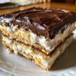 Close-up of a layered slice of eclair cake showing graham crackers, vanilla pudding, whipped cream, and chocolate frosting.