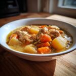 Close-up of a white bowl filled with rich slow cooker chicken stew featuring shredded chicken, large chunks of potato, and bright orange carrots.