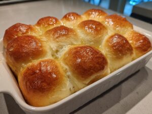 A white baking dish filled with golden brown, glossy sourdough discard rolls brushed with melted butter.
