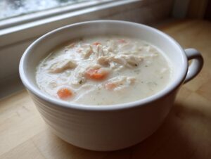 A close-up of a steaming white mug filled with creamy homemade cream of chicken soup, featuring shredded chicken and carrots.