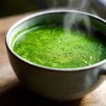 Close-up of a steaming bowl filled with vibrant, creamy spinach soup on a wooden surface.