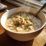 A steaming white bowl filled with creamy white chicken chili, topped with shredded white chicken and green onions.