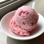Two scoops of bright pink strawberry ice cream with visible fruit pieces served in a small white bowl.