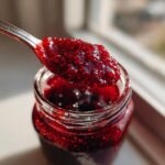A spoonful of vibrant, textured raspberry jam lifted from a glass jar, catching the sunlight.
