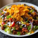 A vibrant, fully loaded taco salad featuring seasoned ground beef, lettuce, tomatoes, olives, and topped with tortilla chips.