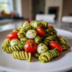 A close-up of a serving of pesto pasta salad featuring rotini pasta, cherry tomatoes, and small mozzarella balls.