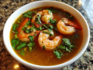 Close-up of a white bowl filled with rich, orange-brown tom yum soup featuring large shrimp and fresh cilantro.