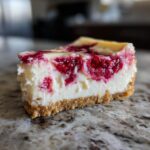 A close-up view of a perfect slice of white chocolate raspberry cheesecake showing the creamy filling and graham cracker crust.