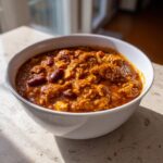 A close-up of a white bowl filled with rich, thick pumpkin chili featuring kidney beans and ground meat.