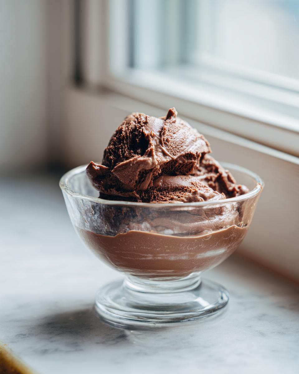 A scoop of rich, dark chocolate mousse served in a small glass dessert dish by a window.